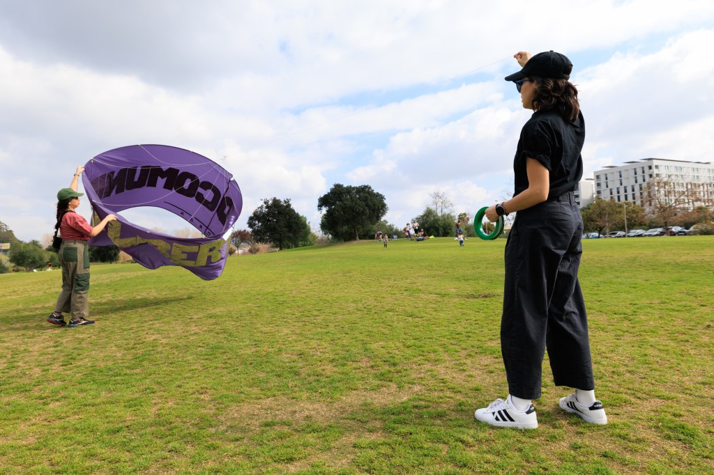 Two people flying a large purple kite in a green field on a cloudy day. One person is holding the kite while the other is managing the string.