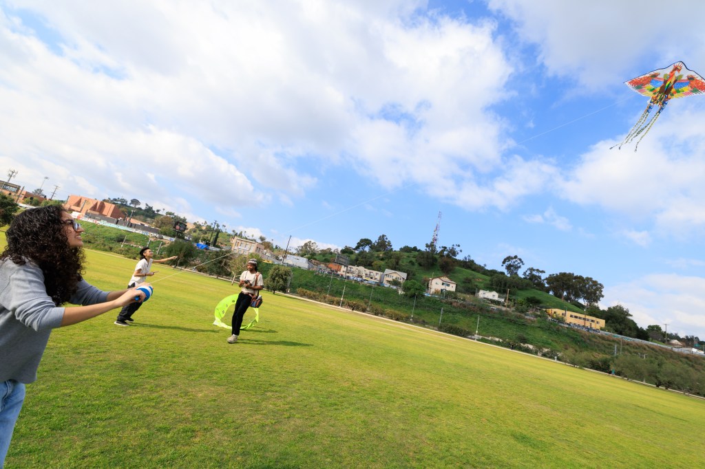 Three people flying a colorful kite on a grassy field under a partly cloudy sky.