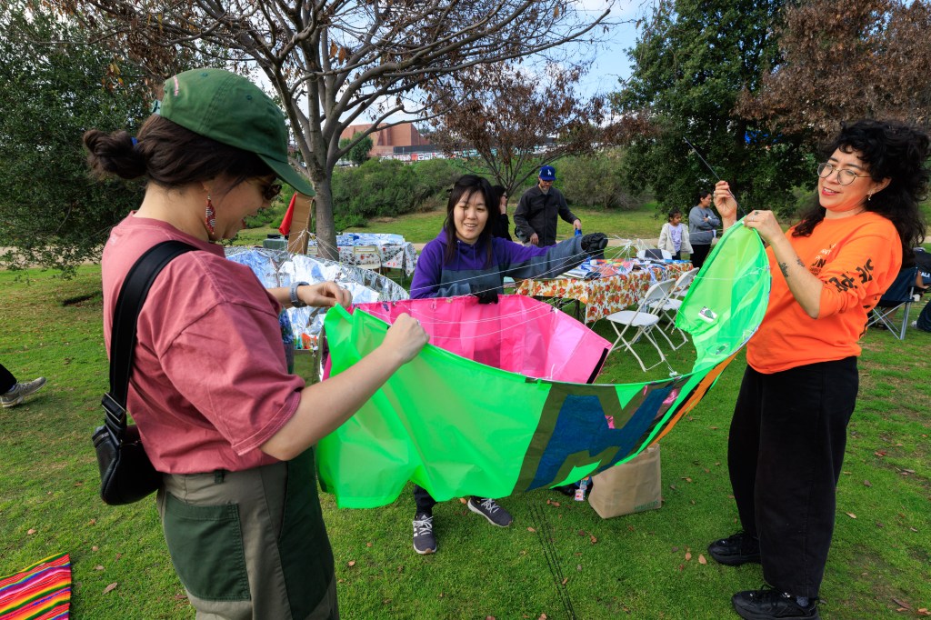 A group of four people outdoors working together to assemble a colorful kite on green grass, with various picnic setups in the background.