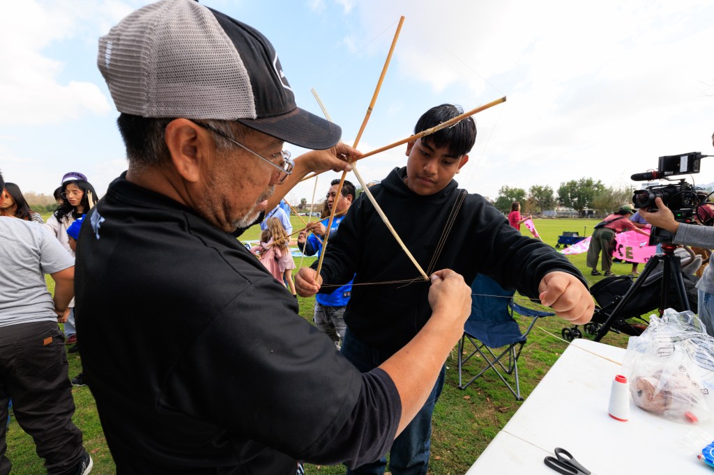 A community event where an older man is teaching a young boy how to make a traditional object using wooden sticks and string. In the background, other participants are engaged in various activities.