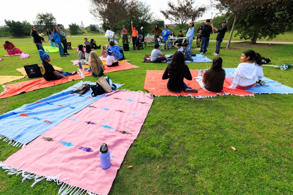 A group of people sitting on colorful blankets in a park, with some engaging in conversation and activities in the background.