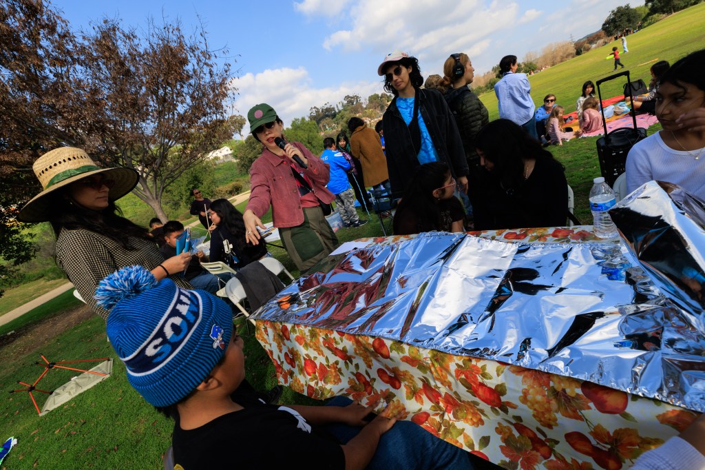 A group of people enjoying a picnic in a park. One person is speaking into a microphone while others listen and engage. A table covered with foil and floral fabric is in the foreground.