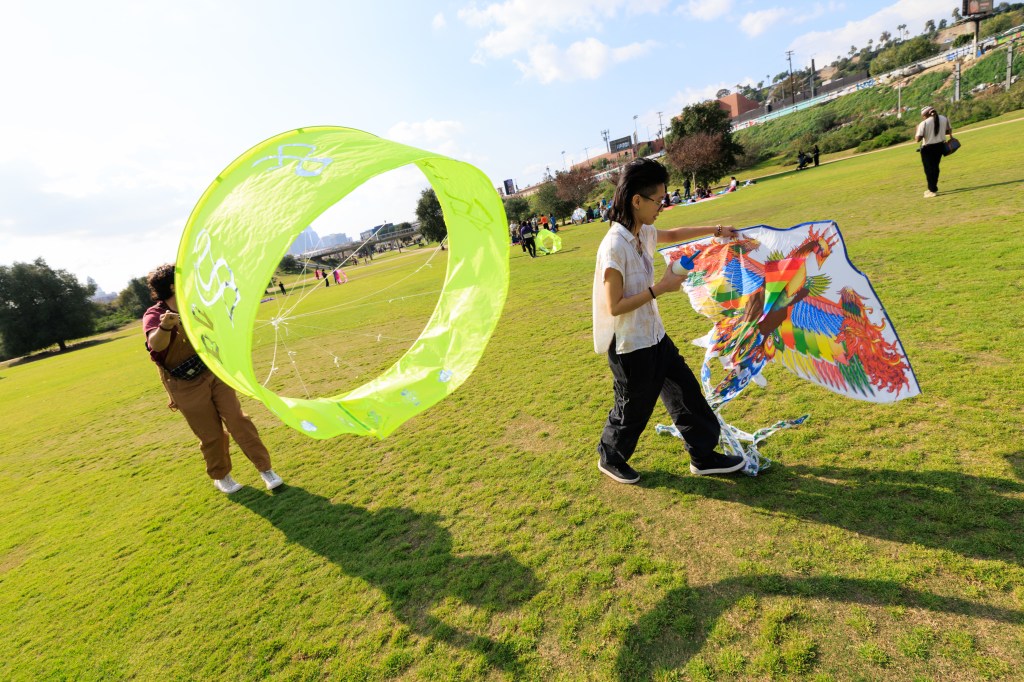 Two people in a park, one holding a large green circular kite and the other carrying a colorful patterned kite.