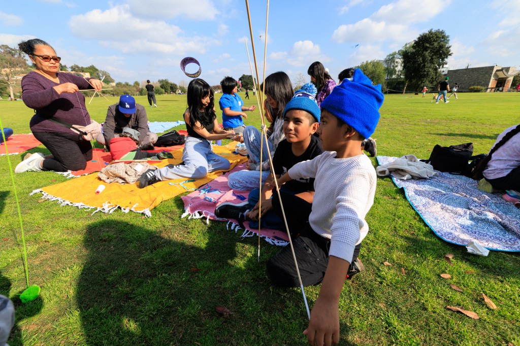 A group of children and adults engage in crafting activities outdoors on colorful blankets in a park setting. One adult is holding a string, while several children are focused on their projects. The scene captures a sunny day with green grass and trees in the background.
